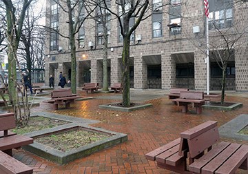 Image of area brick paved plaza with benches and vegetation, with building in background.