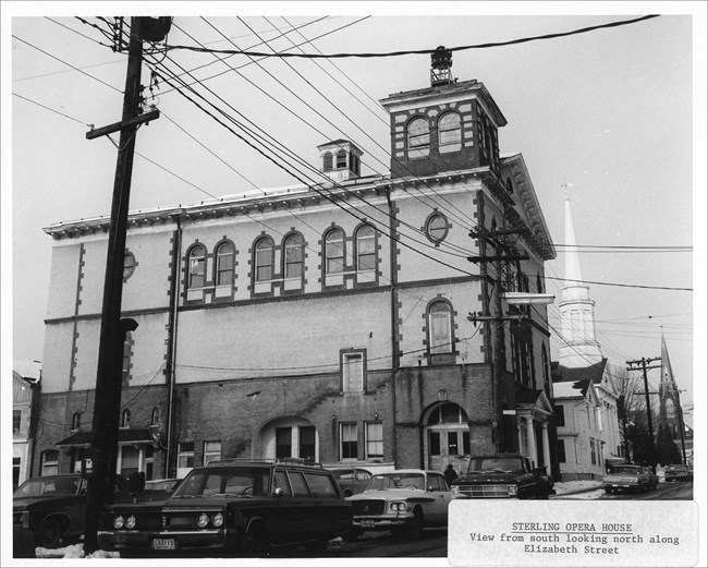 Image of multistory building, cars, road, and electric lines.