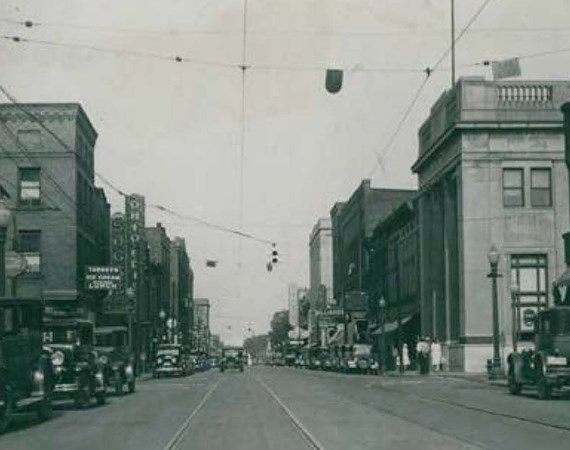 Historic image of street flanked by buildings, with overhead cable lines and cars.