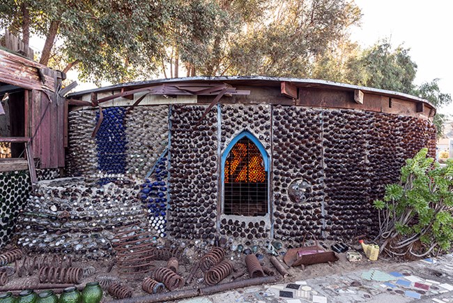 Image of a round single story building constructed of colorful bottle, large metal springs, mosaic walkway, and plants.