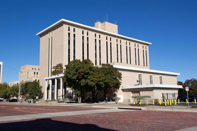 Multi-story light brick rectangular building featuring several columns of narrow windows and an overhanging roof.