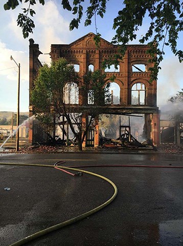 Image of fire-damaged brick building facade, streets, and fire house spraying water.