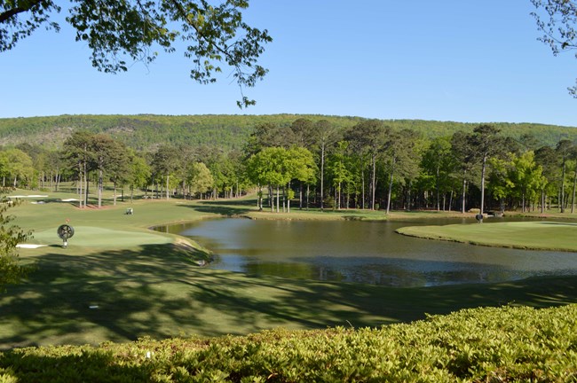 A large golf course beneath a forested hill. A small lake sits within the golf course.