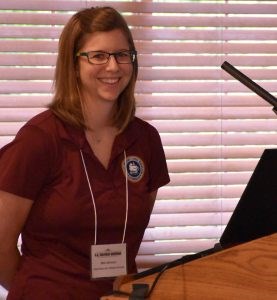 A smiling white woman with long blond hair and glasses wearing a maroon polo shirt and nametag.