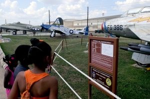 Three black children read a sign about the Republic F-84F Thunderstreak. One silver plane is behind the sign two others are in the background.