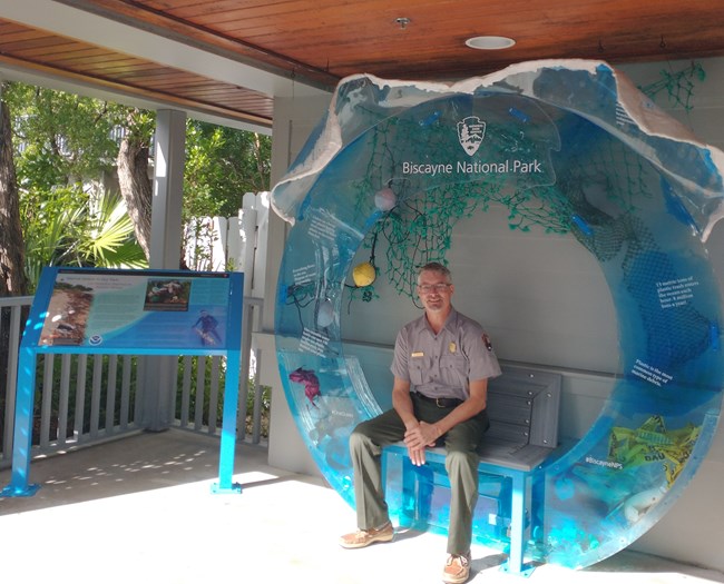 a man in national park service uniform sits on a bench inside a blue acrylic sculpture with ocean trash inside of it. The sculpture sits next to a wayside panel.
