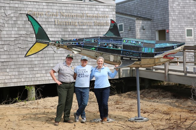 Three smiling women, one in National Park Service Uniform pose for a photo in front of a colorful 14-foot sculpture made out of marine debris items secured to a metal shark frame.