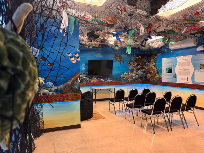 Chairs in a classroom with the walls painted as an underwater scene and pieces of marine debris in nets surrounding the mural.