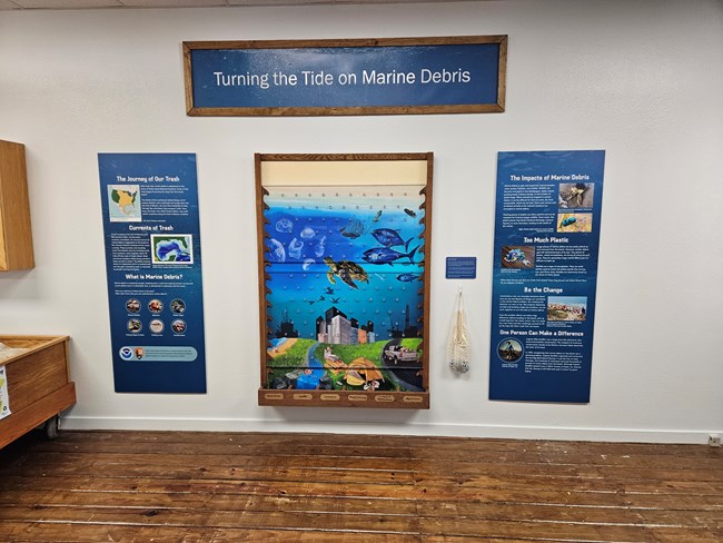Exhibit featuring two educational panels and an interactive Plinko game in the center featuring a painting of how human action interferes with marine life.