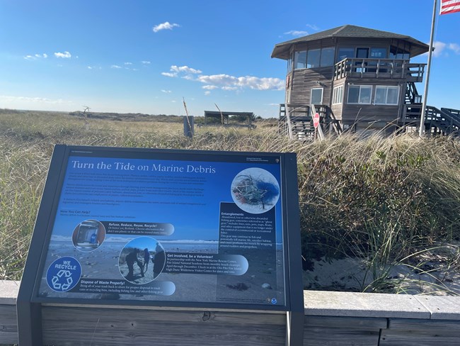 A wayside describing how to “turn the tide on marine debris” on a boardwalk with the Otis Pike Wilderness Visitor Center in the background.