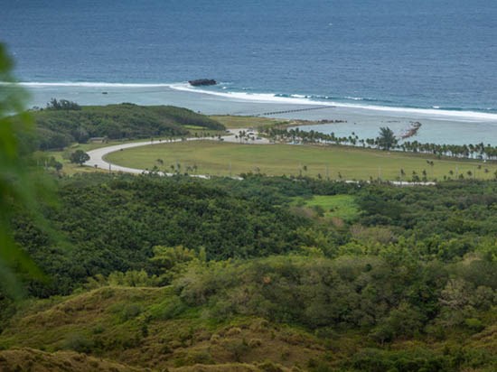 View from elevation over a lush landscape towards a wide area of turf and palms along a bright blue bay.