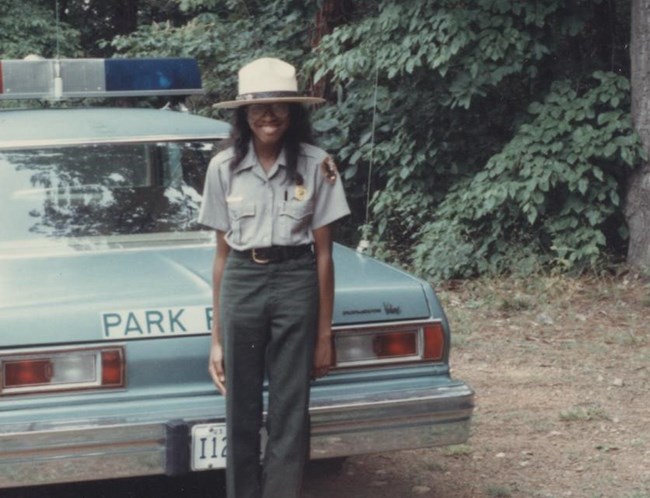 Ajena Rogers stands smiling in NPS ranger uniform and flathat, standing beside a park ranger car beside a wooded area.