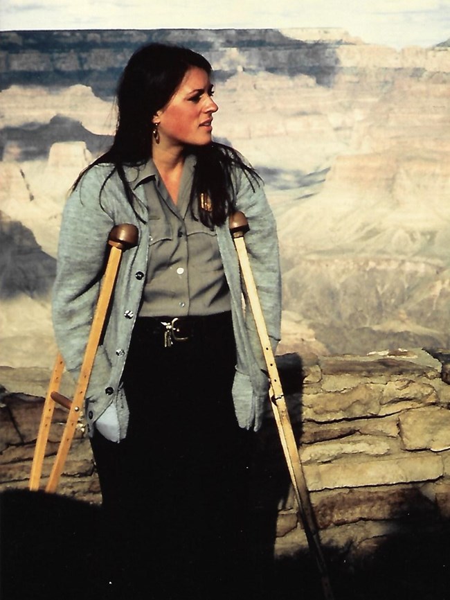 Eileen Szychowski stands near a low stone wall with the Grand Canyon in the background. There are crutches under her arms.