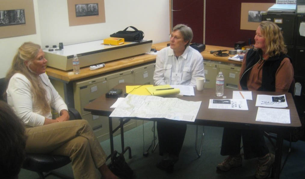 Two people sit at a table with papers, maps, and a recorder to interview a third person, seated to the left.