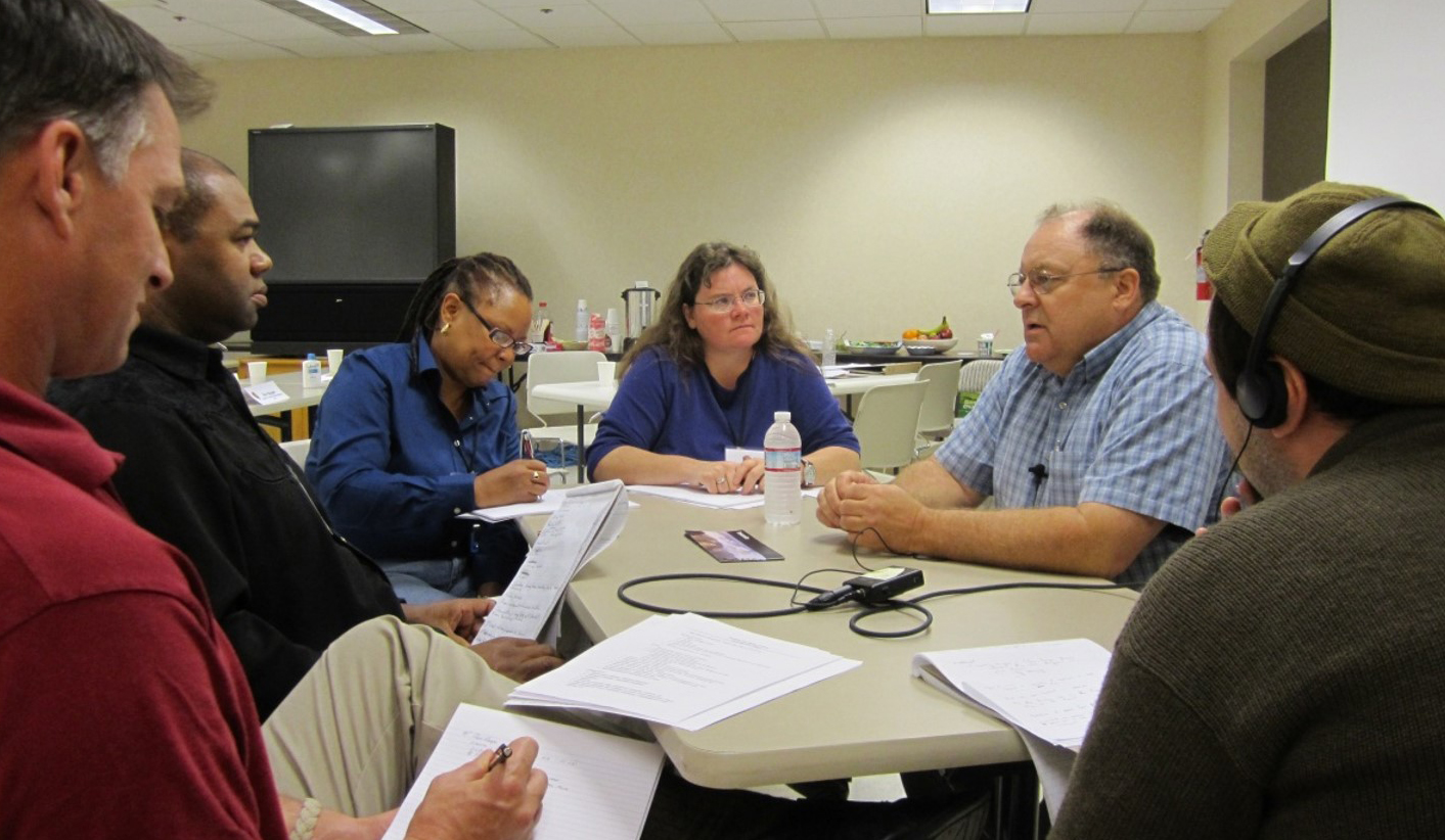 A group of six people seated around a table, with papers, notebooks, and recording equipment in front of them.