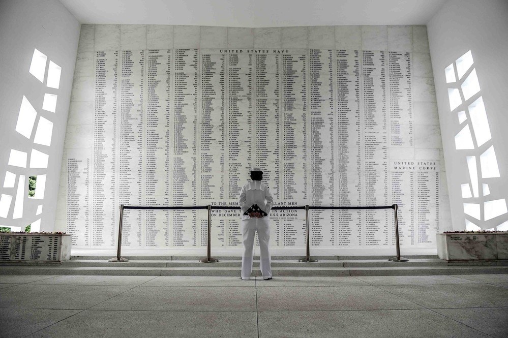 In the Shrine Room at the USS Arizona Memorial, a large wall lists the names of the Arizona’s dead. A person in uniform stands facing the walll, holding a folded American flag.
