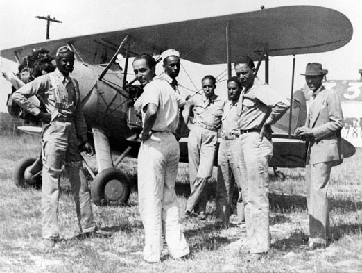 A group of airmen stand near the wing of a biplane in a grassy airfield.