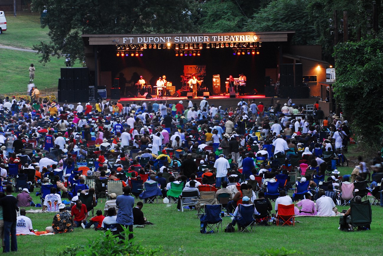 A large crowd is gathered in the grass in front of an outdoor stage that says "Fort Dupont Summer Theater"