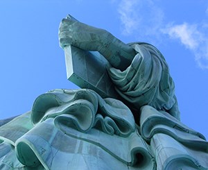 Photo of a part of the Statue of Liberty with Lady Liberty's arm holding a tablet of law above her robe. A blue sky is above her.