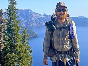 a smiling man with a red beard wearing a backpack stands in front of a blue lake with mountains in the distance