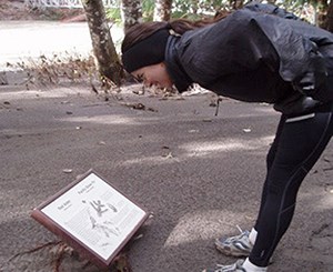 A woman wearing sneakers, black tights, gray jacket and black headband bends over to read a sign almost buried in mud. Behind her are trees, a river and a bridge.