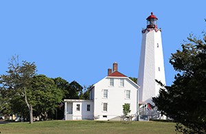 White lighthouse with a red bell tower next to a white house with a red roof on a green lawn next to green trees with a blue sky