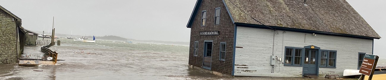 A small wooden building covered in shingles is affected by storm tides. The water completely encroaches on the base, and reaches up the road.