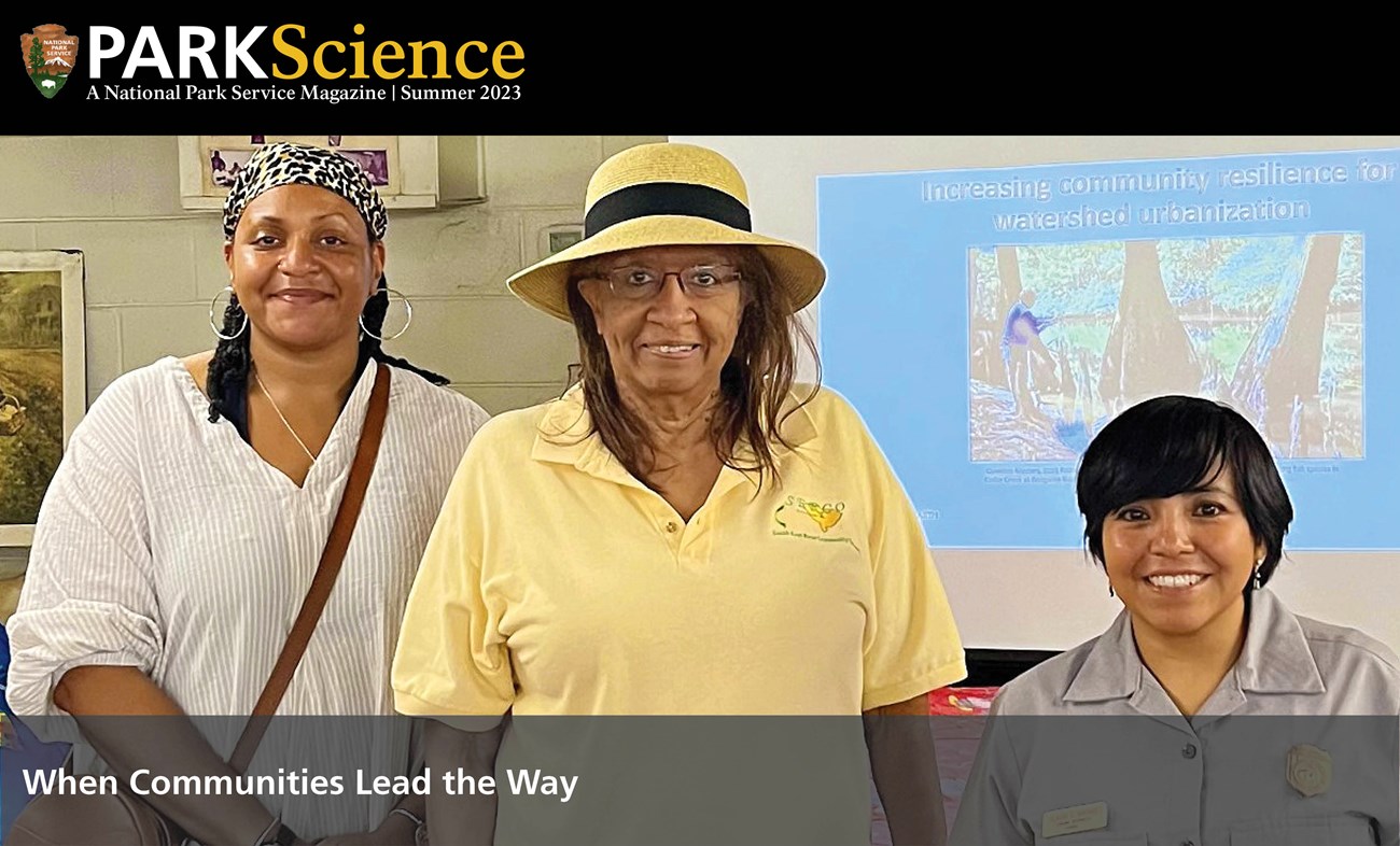 Three smiling women standing in front of a wall with decorations and a slide show. One is wearing an NPS uniform.