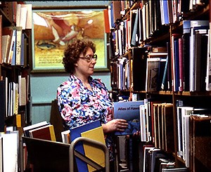 A woman with glasses in bright shirt with flower pattern holds a book as she returns it to a library shelf. She is standing in front of a picture on the wall and between two tall shelves of books, A library cart is in front of her.