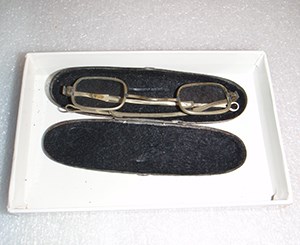 A pair of old eyeglasses in a pale metal frame in an oval glasses case with a black interior.