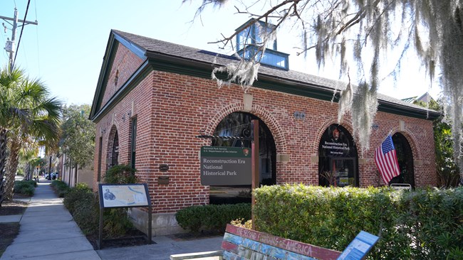 A brick single story building with large arched windows and trees surrounding the exterior.