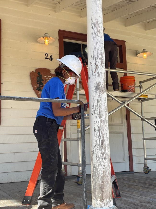 A woman wearing personal protection equipment works on scraping paint off a column on the porch of Darrah Hall.