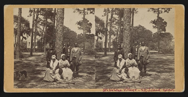 Three men stand, two women, and a child sit outside in front of pine trees and houses.