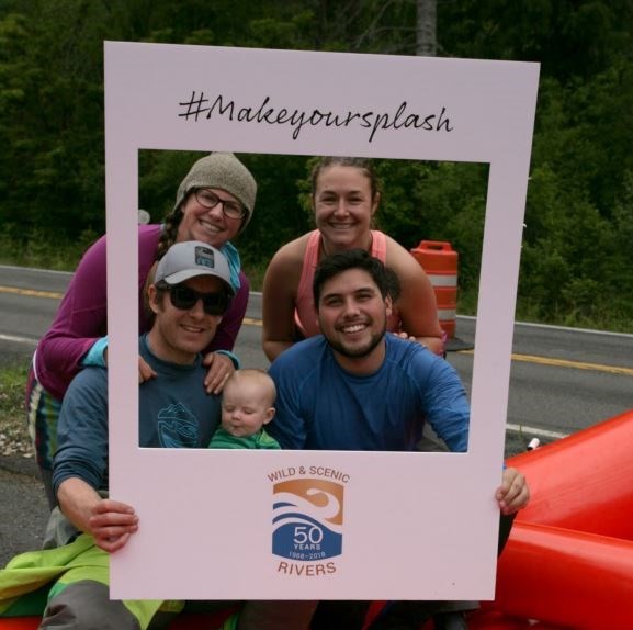 Four adults and a baby sit in a raft on the side of the road, smiling while holding a cardboard #Makeyoursplash photo frame around them.