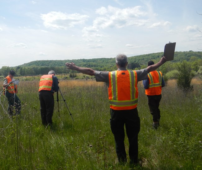 A team of 4, evaluate a view at Delaware Water Gap National Recreation Area. A man stands with his both arms out to show the boundaries of the view. On man looks into a camera on a tripod. Other team members are looking at the view of a hill.