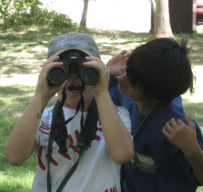 A boy looks through binoculars with gaping mouth as a second boy tries to look over his shoulder.