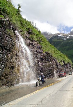 A motorcycle and two automobiles drive past a waterfall on a scenic mountain road.