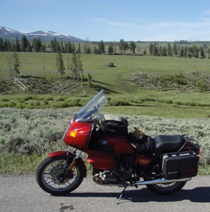 Red motorcycle parked on roadside with meadow, trees, and mountains in background.