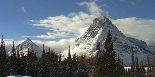 Two snowy mountain peaks, with an avalanche rushing down the side of the larger one. Evergreen trees in the foreground.