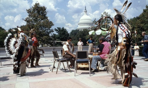 Men in Omaha Indian regalia dance while others play drums, with the Capitol building in the background.