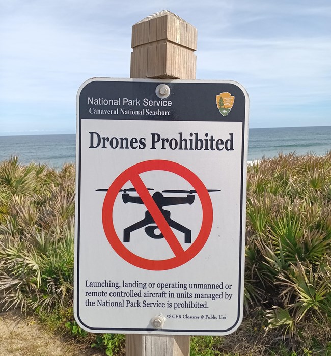 Photo of a sign reading "Drones Prohibited: Launching, landing, or operating unmanned or remote controlled aircraft in units managed by the National Park Service is prohibited". The sign also features an icon of a drone crossed out. Ocean in background