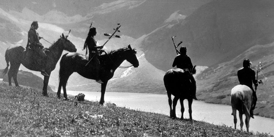 Historic black and white photo of four Native Americans sitting on horses with a mountain and mountain lake in the distance.
