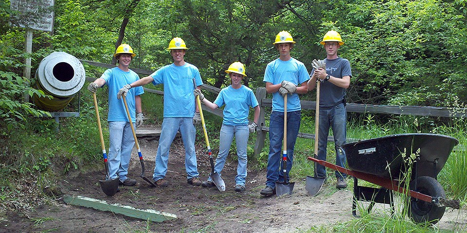 Five youth wearing blue t-shirts and yellow hard hats hold yellow-handled shovels next to a black wheelbarrow.