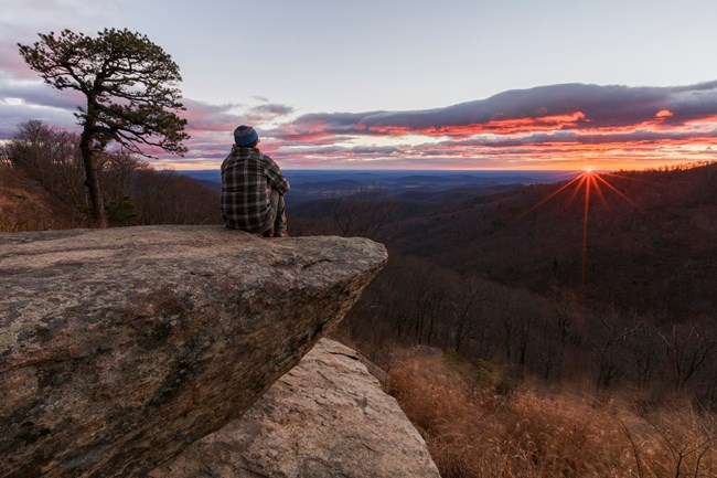 a person sits on a rocky ledge watching the sunset over rolling hills