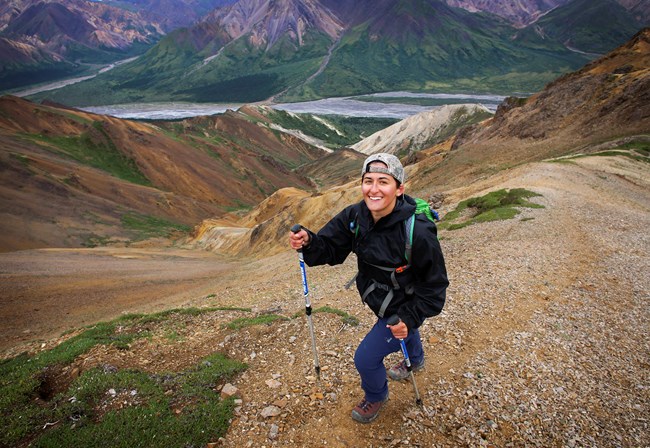 a hiker with hiking poles smiles at the camera while ascending a steep mountain slope