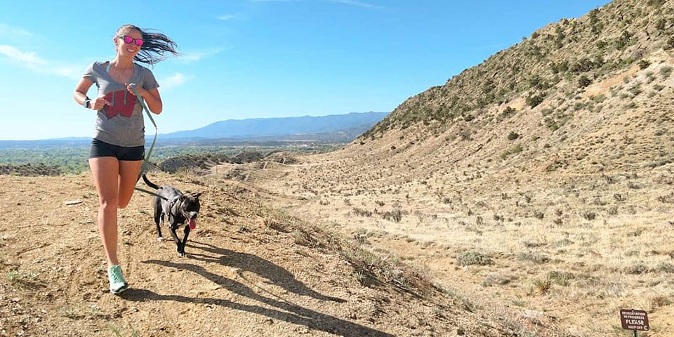A woman and leashed dog run on a dirt trail