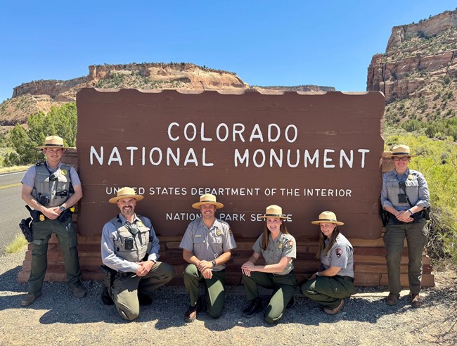 Six people stand in front of the Monument's entrance sign wearing NPS uniforms.