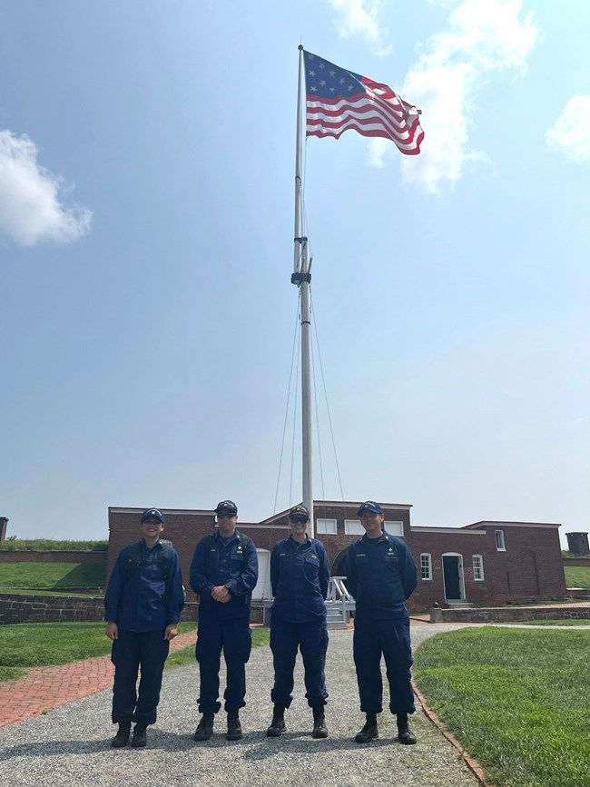 Four staff are standing in front of a flag pole wearing blue uniforms.