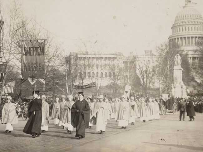 Group of white female nurses in early 20th century nursing outfits marching in front of the Capitol building