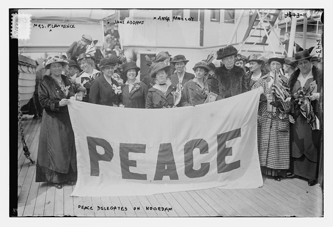 A group of women stand on the deck of a ship holding a large sign that says PEACE